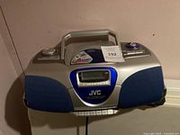 Front view of JVC boombox showing silver and blue casing, handle, and speaker grills.