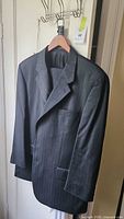 Front view of men's dark wool suit jacket with pinstripe pattern hanging on a wooden hanger against a white door.