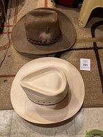 Two men's cowboy hats side by side: brown suede hat with feather-accented band and white straw Resistol hat.