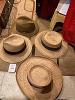 Four natural-colored straw men's summer hats arranged showing tops and sides, each with a different decorative band.