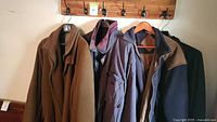 Four men's coats hanging on wall hooks: camel wool coat, purple insulated jacket, navy blue fleece jacket with brown shoulders, dark coat.
