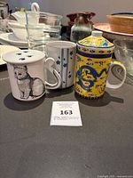 Three unique mugs displayed on a table, front view showing cat mug with paw print lid, tall floral striped mug and yellow dragon mug.
