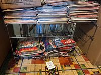 Photo shows multiple stacks of vintage magazines on a metal rack with some magazines on the floor beneath the rack. Covers include automotive and entertainment themes.