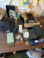 Photo showing various office desk supplies on a wooden desk including a vintage calculator, metal box containers, a blue tape dispenser, and a coin or medallion.