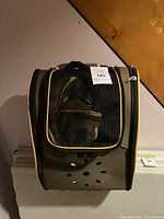 Photo of a small animal carrier designed for XS dogs or small cats. The carrier is gray with a black mesh front panel outlined in yellow piping. Several ventilation holes are visible. The background includes a wooden wall and the carrier is hanging or placed against a wall.