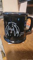 Photo of black ceramic mug displaying white minimalist line drawing of John Lennon with signature and years 1940-1980.