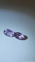 Pair of oval cut pink-purple amethyst gemstones placed on white surface, showing color and cut details.