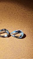 Close-up of two pear-shaped aquamarine gemstones on a textured brown surface showing reflections and internal clarity details.