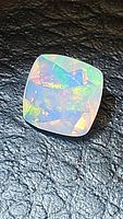 Close-up photo of a cushion cut Ethiopian opal faceted gemstone on a textured black background. The stone shows clear faceting and bright rainbow color flashes.