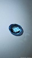 Close-up image of a natural Swiss blue topaz gemstone, oval checkerboard cut, deep lagoon blue color, placed on a white background.
