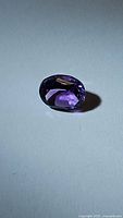 Close-up front view of the deep purple, faceted oval amethyst gemstone on a white background to show color and cut details.