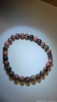 Natural rhodonite bead bracelet laid flat in circular shape on white surface, showing smooth polished beads with mixed pink and black colors.