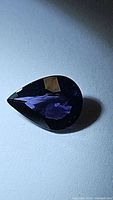 Pear-shaped purple spinel gemstone with dark rich purple color and reflective facets, photographed on white surface to show cut and clarity.
