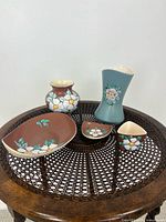 Five BC Ceramics pieces arranged on a round wooden table with brown woven top showing various dogwood and wild rose floral designs