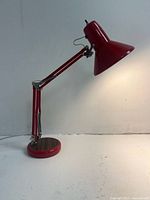 Red vintage style articulated table lamp shown fully extended showing adjustable arm and lampshade with heavy round base on white surface against white background.