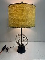 Front view of the mid-century modern globe lamp showing the fiberglass cylindrical shade with yellowish woven texture and black stitched trim, along with the metal globe base on a black conical stand, electric cord and pull switch.