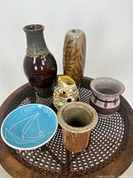 Image showing six ceramic studio pottery items on a metal mesh and wood circular tray. Includes variously sized vases, a small owl figurine, and a decorative plate with sailboat design.