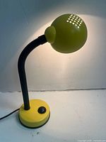 Photo of a yellow gooseneck desk lamp turned on, showing perforated top on round head and flexible black arm attached to round yellow base with black switch.