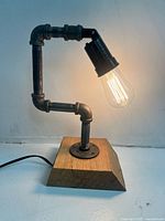 Photo of a steampunk industrial pipe table lamp with Edison bulb and wooden base, lit and shown against a plain wall.