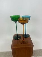 Set of three colored glass compotes displayed on a wooden furniture piece, showing green, amber, and blue compotes with textured floral patterns