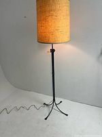 Full view of lamp illuminated, showing cylindrical beige fabric shade and wrought iron shaft with tripod base