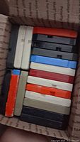 Photo of a box filled with 15 assorted 8-track cartridges in various colors including black, white, red, blue, and gray, small visible wear on edges.
