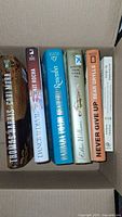 Seven books arranged standing upright in a cardboard box viewed from above, showing spines and some front covers.