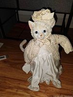 Front view of the porcelain cat doll showing detailed painted face, blue eyes, knitted sweater, creamy white silky dress and decorative hat.