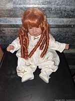 Front view of porcelain doll sitting, showing red curly hair and white outfit with gold accents.