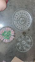 Three decorative glass serving plates arranged on a dark floor, showing variety in design including clear textured, clear floral starburst, and pink floral hand-painted plate.