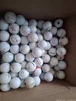 Golf balls inside a cardboard box, mostly white with various branding and colored stripes or logos visible.