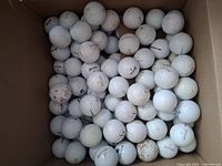 Photo showing a cardboard box filled with approximately 100 white golf balls of various brands with scuffs, dirt, and stains.