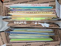 Side view of stacked greeting cards and envelopes in a USPS shipping box showing the thickness and quantity of the lot.