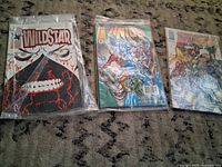 Three comic books displayed on carpet: WildStar issue, another issue with energy blast and skeleton face, and a group battle scene issue. Each individually bagged.