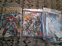 Three comic books laid out side by side showing cover art including Brigade and other superhero titles in plastic sleeves.