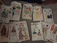 Wide view of assorted vintage sewing pattern envelopes laid out on a carpet. Various brands and clothing styles represented, including Butterick, Simplicity, and Advance. Patterns show women and children's fashion items from around the 1950s-1970s era.