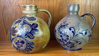 Photo showing two round-bodied German stoneware pitchers with incised blue floral and wheat decorations and handles, placed on a wooden surface against wooden backdrop.