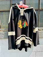 Front view of black hooded sweatshirt with cream lace-up neckline and cream striping on sleeves and bottom hem, featuring Chicago Blackhawks logo.