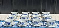 Twelve tea cups and saucers arranged in four rows of three on a woven tablecloth.