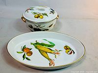 Photo showing oval serving dish decorated with corn, apples, and other fruits, alongside lidded casserole dish with lemon motifs and gold trim.