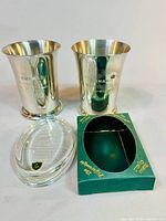 Two polished silver-tone pewter cups engraved with Cunard World Cruise and a clear Stuart crystal oval paperweight in a green box.