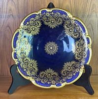 Front view of the vintage cobalt blue cabinet plate with detailed gold gild pattern and scalloped edges, displayed on a black stand.