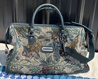 Front side of a rectangular travel bag with floral tapestry design, black leather handles, and a black shoulder strap under natural light.