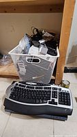 Basket of assorted electronics and silver/black keyboard on floor