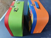 Side view of two metal puzzle carrier cases, one green and red, the other blue and orange, showing metal clasps and handles, one handle missing.