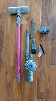 Dyson V6 vacuum and attachments laid out on wooden floor, showing pink extension wand, various nozzle and brush attachments