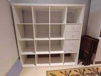 Straight-on photo of white Ikea KALLAX shelving unit with 16 compartments and 4 drawers on right side.