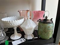 Shelf view showing assorted glassware including milk glass compote, octagonal covered dish, two pink glass pitchers, green biscuit jar