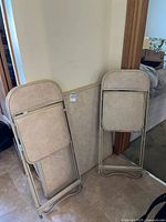 Two beige metal folding chairs leaning against wall beside folded card table top