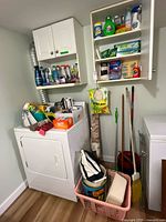 Wide view of laundry area showing vacuum, broom, mop, laundry basket and upper cabinets with supplies
